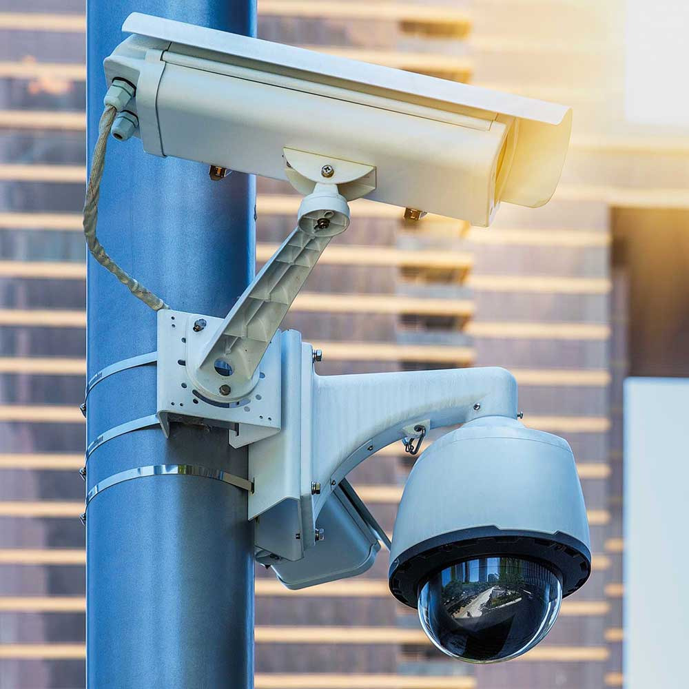 Two cameras on a pole in a public space. The sun shines on the camera from the top right.