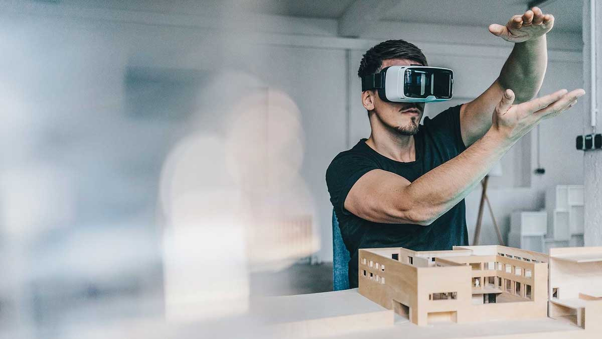 A man wears virtual reality glasses and gestures with his hands. In front of him is a model of a building.